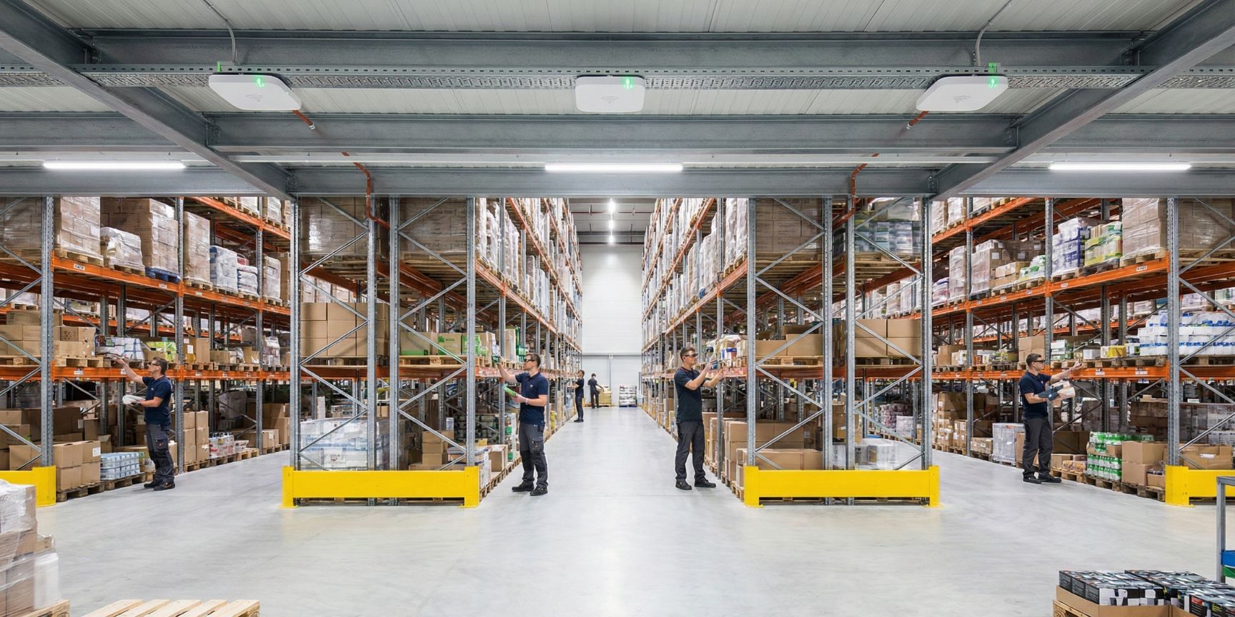 Weitwinkelaufnahme einer modernen Hochregallagerhalle mit mehreren Mitarbeitern, die Datenbrillen zur Kommissionierung tragen. An der Hallendecke sind deutlich mehrere WLAN Access Points für eine flächendeckende Ausleuchtung installiert